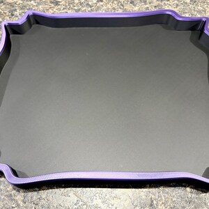 Pode incluir: Uma bandeja quadrada com uma base interior em preto mate e uma borda elevada e ondulada em roxo. A bandeja apresenta um design distinto com bordas recortadas e ornamentadas que emolduram a superf&iacute;cie central plana e escura.