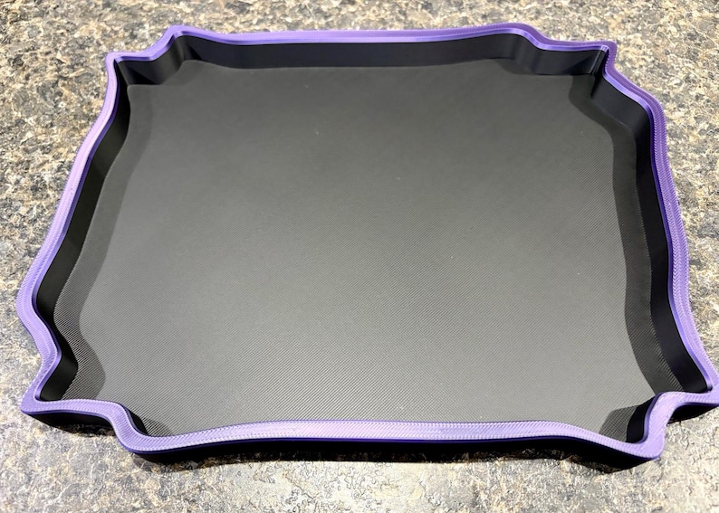 Pode incluir: Uma bandeja quadrada com uma base interior em preto mate e uma borda elevada e ondulada em roxo. A bandeja apresenta um design distinto com bordas recortadas e ornamentadas que emolduram a superf&iacute;cie central plana e escura.