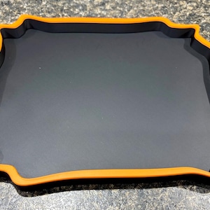Pode incluir: Uma bandeja quadrada com uma superf&iacute;cie interior preta fosca e uma borda elevada em laranja vibrante. A bandeja apresenta um design distinto de bordas onduladas que cria uma apar&ecirc;ncia de moldura ao redor da base central plana e escura.