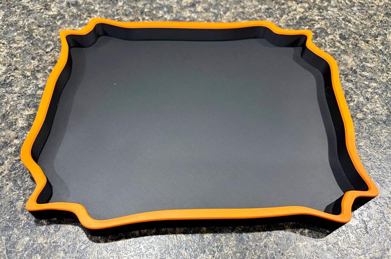 Pode incluir: Uma bandeja quadrada com uma superf&iacute;cie interior preta fosca e uma borda elevada em laranja vibrante. A bandeja apresenta um design distinto de bordas onduladas que cria uma apar&ecirc;ncia de moldura ao redor da base central plana e escura.