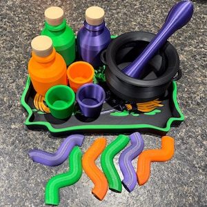 Pode incluir: Um conjunto de acess&oacute;rios para po&ccedil;&otilde;es, incluindo um caldeir&atilde;o preto com um pil&atilde;o roxo, duas garrafas com rolhas bege, tr&ecirc;s copos pequenos nas cores laranja, verde e roxo, um tabuleiro preto com gr&aacute;fico de vassoura e seis formas onduladas.