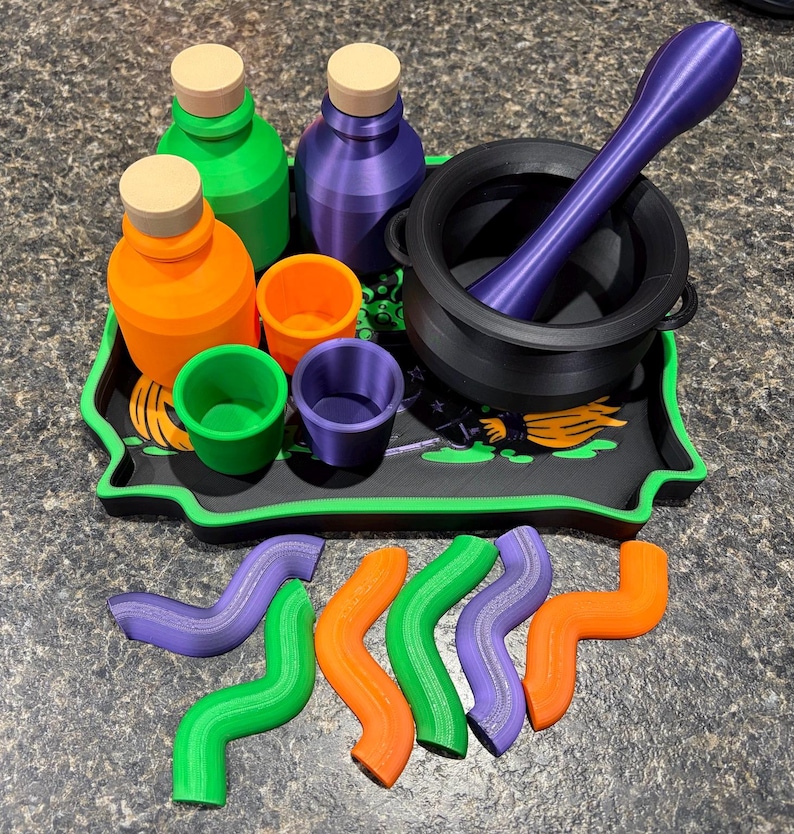 Pode incluir: Um conjunto de acess&oacute;rios para po&ccedil;&otilde;es, incluindo um caldeir&atilde;o preto com um pil&atilde;o roxo, duas garrafas com rolhas bege, tr&ecirc;s copos pequenos nas cores laranja, verde e roxo, um tabuleiro preto com gr&aacute;fico de vassoura e seis formas onduladas.