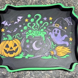 Pode incluir: Uma bandeja retangular preta com uma borda verde brilhante apresentando um design colorido de Halloween. O gr&aacute;fico inclui uma ab&oacute;bora laranja, um caldeir&atilde;o verde borbulhante com uma lua crescente, um fantasma roxo, uma vassoura e morcegos.