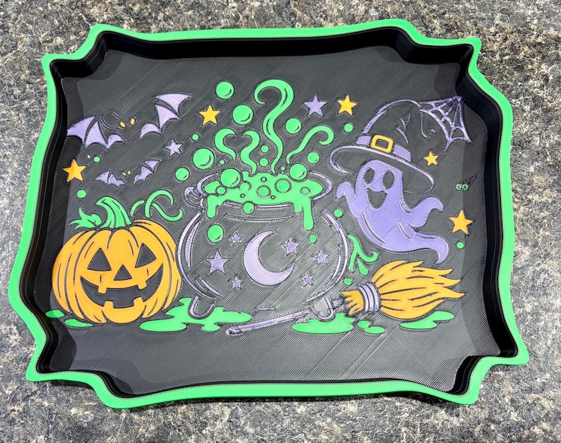 Pode incluir: Uma bandeja retangular preta com uma borda verde brilhante apresentando um design colorido de Halloween. O gr&aacute;fico inclui uma ab&oacute;bora laranja, um caldeir&atilde;o verde borbulhante com uma lua crescente, um fantasma roxo, uma vassoura e morcegos.