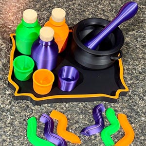 Pode incluir: Uma bandeja preta com borda laranja cont&eacute;m tr&ecirc;s garrafas com rolhas bege, tr&ecirc;s copos pequenos e um caldeir&atilde;o preto com um pil&atilde;o roxo. As garrafas e copos s&atilde;o verdes, laranja e roxos. Seis formas onduladas e coloridas est&atilde;o dispostas perto.