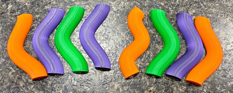Pode incluir: Uma cole&ccedil;&atilde;o de oito objetos tubulares curvos dispostos em uma fileira. Os itens apresentam uma forma ondulada e serpentina distinta e s&atilde;o coloridos em tons alternados de laranja brilhante, roxo vibrante e verde lim&atilde;o com textura.