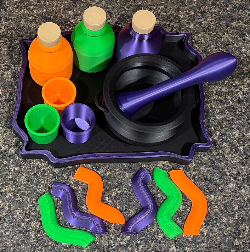 Pode incluir: Uma bandeja preta cont&eacute;m tr&ecirc;s garrafas com rolhas bege em laranja, verde e roxo, tr&ecirc;s copos a condizer e um almofariz com pil&atilde;o preto. Seis formas onduladas em laranja, verde e roxo est&atilde;o dispostas &agrave; frente da bandeja.