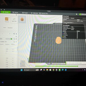 Pode incluir: Um ecr&atilde; de computador a exibir a interface do software Bambu Studio para impress&atilde;o 3D. O ecr&atilde; mostra uma pr&eacute;-visualiza&ccedil;&atilde;o digital de um pequeno objeto cil&iacute;ndrico laranja centrado numa placa de constru&ccedil;&atilde;o de grelha preta com dados.