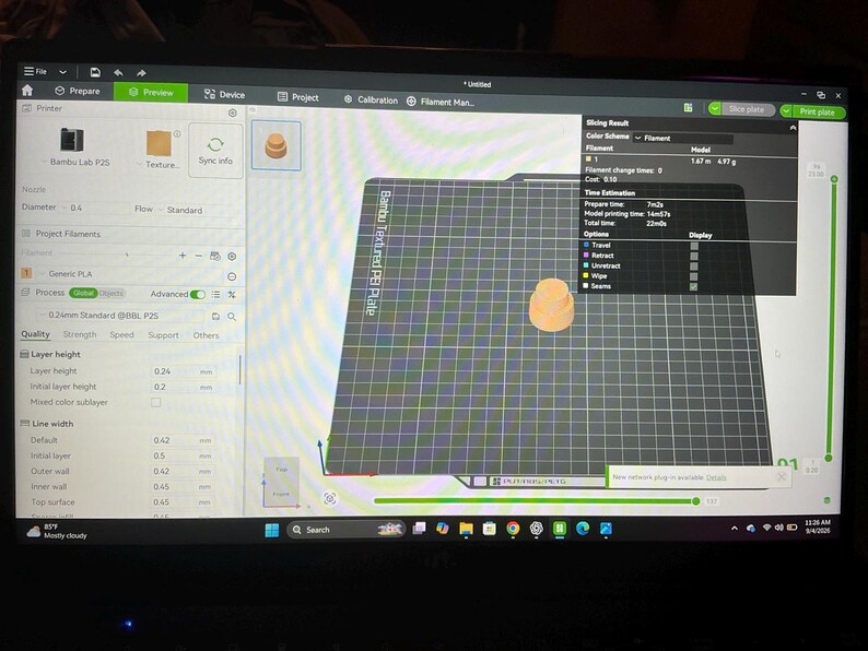 Pode incluir: Um ecr&atilde; de computador a exibir a interface do software Bambu Studio para impress&atilde;o 3D. O ecr&atilde; mostra uma pr&eacute;-visualiza&ccedil;&atilde;o digital de um pequeno objeto cil&iacute;ndrico laranja centrado numa placa de constru&ccedil;&atilde;o de grelha preta com dados.