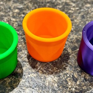 Pode incluir: Tr&ecirc;s pequenos recipientes cil&iacute;ndricos de topo aberto em cores s&oacute;lidas vibrantes. Um recipiente &eacute; verde vibrante, um &eacute; laranja brilhante e um &eacute; roxo profundo. Cada copo apresenta uma borda lisa e uniforme e um design interno oco.