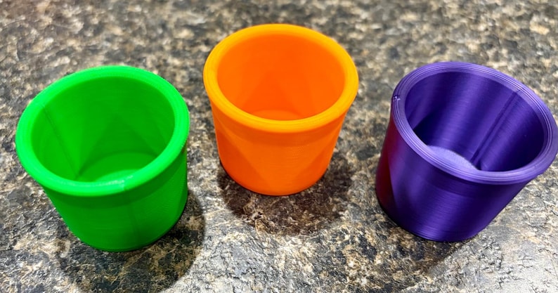 Pode incluir: Tr&ecirc;s pequenos recipientes cil&iacute;ndricos de topo aberto em cores s&oacute;lidas vibrantes. Um recipiente &eacute; verde vibrante, um &eacute; laranja brilhante e um &eacute; roxo profundo. Cada copo apresenta uma borda lisa e uniforme e um design interno oco.