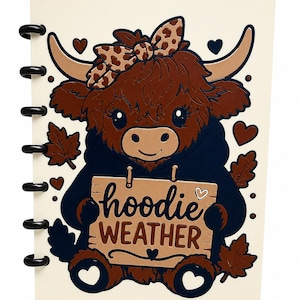 Könnte beinhalten: Ein cremefarbenes Notizbuch mit Scheibenbindung, das ein braunes Hochlandrind mit dunkelblauem Kapuzenpullover und einer Schleife mit Leopardenmuster zeigt. Das Rind hält ein Holzschild mit der Aufschrift "hoodie WEATHER" und herbstlichen Blättern.