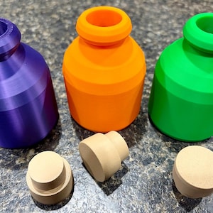 Pode incluir: Tr&ecirc;s frascos pequenos de boca larga nas cores roxa, laranja e verde brilhante, cada um com um corpo liso e arredondado e uma borda espessa. Tr&ecirc;s rolhas cil&iacute;ndricas separadas, de cor bege claro e design escalonado, est&atilde;o &agrave; frente dos frascos.