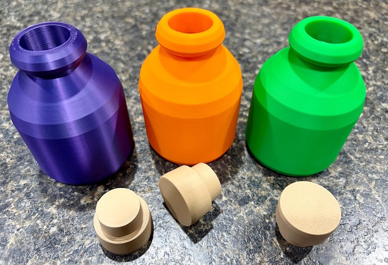 Pode incluir: Tr&ecirc;s frascos pequenos de boca larga nas cores roxa, laranja e verde brilhante, cada um com um corpo liso e arredondado e uma borda espessa. Tr&ecirc;s rolhas cil&iacute;ndricas separadas, de cor bege claro e design escalonado, est&atilde;o &agrave; frente dos frascos.