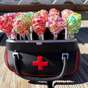 May include: A black plastic container shaped like a medical bag with a red cross emblem on the front, filled with multiple Dum Dums lollipops in various colorful wrappers including strawberry, mystery, and cream soda flavors.