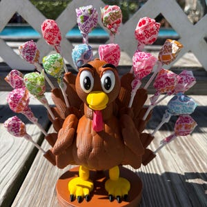 May include: A brown 3D-printed turkey figurine with a yellow beak and red wattle, standing on a circular base. The turkey features slots in its back holding multiple Dum-Dums lollipops in various colorful wrappers, creating a fan-like tail display.