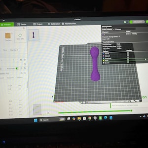 Pode incluir: Uma tela de computador exibindo a interface do software Bambu Studio com um modelo 3D de um objeto roxo em forma de haltere centralizado em uma placa de constru&ccedil;&atilde;o digital. O software mostra um tempo total de impress&atilde;o de 1h 20m.
