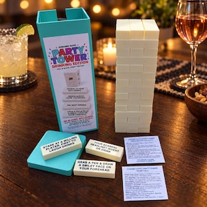 May include: A Kokomo Kove Party Tower drinking game set featuring a teal box, a tall stack of white rectangular wooden blocks, and two instruction cards. Several blocks are printed with text prompts like Stare at a willing player.