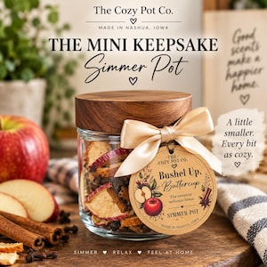 May include: A clear glass jar with a dark wood lid, filled with dried apple slices and cinnamon sticks. A beige satin ribbon ties a round paper tag reading 'Bushel Up, Buttercup' and 'The Cozy Pot Co.' to the jar neck. Text includes 'The Mini Keepsake Simmer Pot'.