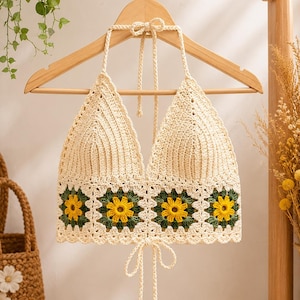 Puede incluir: Un top halter de ganchillo color crema con un corpiño triangular acanalado y una banda inferior compuesta por cuatro motivos cuadrados. Cada cuadrado muestra una flor amarilla con pétalos verdes sobre fondo crema y cierre de lazo.