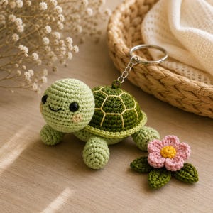 May include: A crochet turtle keychain featuring a light green head with black eyes and a stitched smile, a dark green shell with yellow detailing, and a small pink flower with a yellow center and green leaves attached to the side by a metal ring.