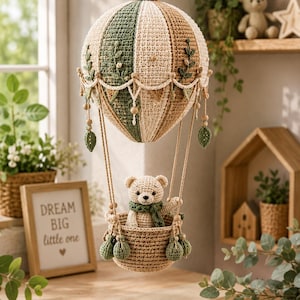 Puede incluir: Un móvil de globo aerostático tejido a ganchillo con una cúpula a rayas en beige, verde salvia y tostado, decorada con hojas bordadas. Un pequeño oso de peluche tejido lleva una bufanda verde y está sentado en la cesta de ganchillo.