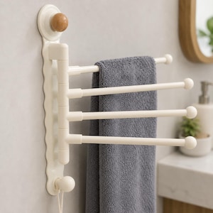 Puede incluir: Un toallero de pared en color blanco roto con cuatro brazos horizontales giratorios con extremos redondeados. Un pomo de color madera se sitúa en la parte superior y otro a juego en color blanco roto en la inferior. Una toalla gris cuelga.
