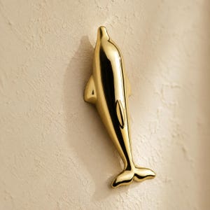 May include: A polished gold-toned dolphin figurine mounted on a wall. The dolphin is depicted in a vertical orientation with a smooth, reflective surface that catches the light, showing a streamlined body shape with a defined snout and tail fin.