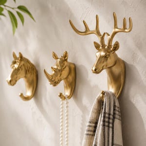 May include: Three gold-coloured wall hooks shaped like animal heads, including a horse, a rhinoceros, and a deer with large antlers. The deer hook holds a plaid fabric, and the rhinoceros hook holds a string of white beads, providing functional storage.