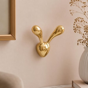Puede incluir: Un gancho de pared de color dorado con una superficie lisa y reflectante, con un diseño en forma de Y que presenta dos puntas redondeadas orientadas hacia arriba que se extienden desde una base central. El objeto metálico está en una pared.