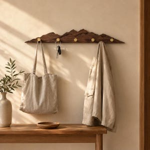 May include: A dark brown wooden wall-mounted coat rack shaped like a mountain range with five circular gold-toned metal hooks. The rack holds a beige canvas tote bag, a set of keys on a ring, and a light tan jacket hanging from the hooks.