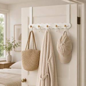 May include: A white over-the-door hook rack featuring a scalloped edge design and five hooks with natural wood ball tips. The rack is mounted on a door and holds a woven straw tote bag, a beige waffle-knit robe, and a beige baseball cap.