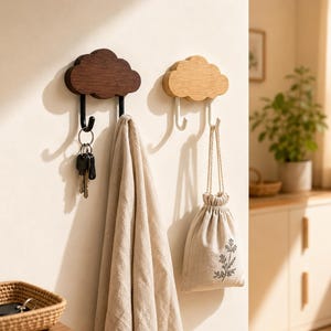 May include: Two cloud-shaped wall hooks made of wood. The left hook is dark brown with a black metal hanger holding a set of keys. The right hook is light tan with a white metal hanger holding a small beige drawstring bag. A beige cloth hangs below.