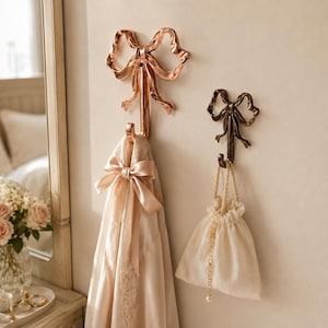 May include: Two wall-mounted hooks shaped like bows. The larger hook is metallic rose gold and holds a light pink satin garment with a matching ribbon. The smaller hook is dark bronze and holds a small, cream-colored fabric drawstring pouch.