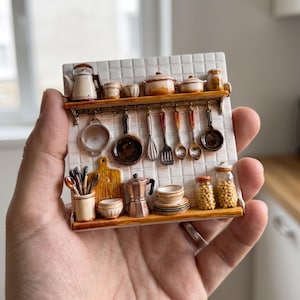 Puede incluir: Una placa de pared de cerámica en miniatura que muestra un salpicadero de cocina de azulejos blancos con dos estantes de madera. Incluye utensilios de cocina diminutos como una tetera, ollas, sartenes, un batidor, una espátula y frascos.