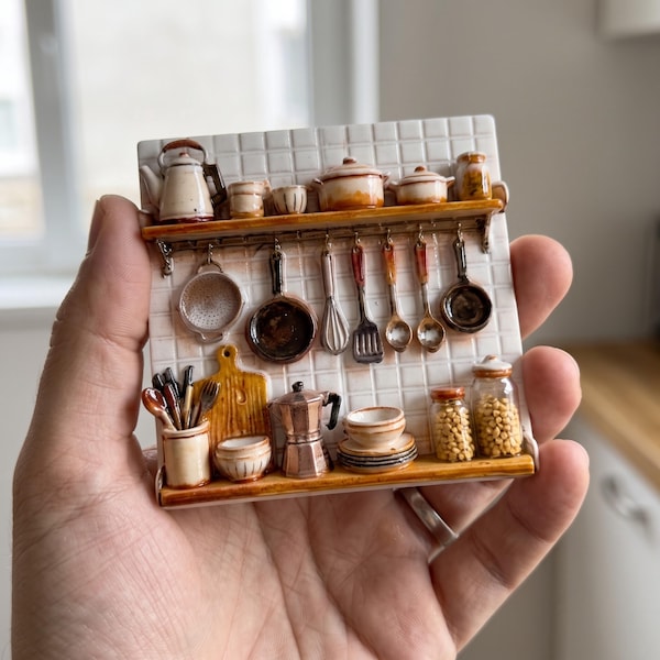 May include: A miniature ceramic wall plaque depicting a kitchen scene with white tiled walls, two wooden shelves, and various kitchenware including a kettle, pots, pans, utensils, a cutting board, a moka pot, and glass jars filled with grains.
