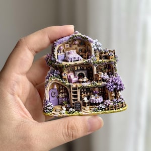 May include: A miniature three-story house figurine featuring purple floral accents, a lavender door, and detailed interior rooms including a bedroom, living area, and kitchen. The structure is adorned with climbing purple flowers and greenery.