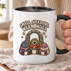 May include: A white ceramic mug with a black handle and rim, featuring a graphic of three small, colourful creatures wearing scarves and holding tiny mugs under a stone archway, with the text "SMALL SHADOWS, WARM MUGS." and yellow stars.