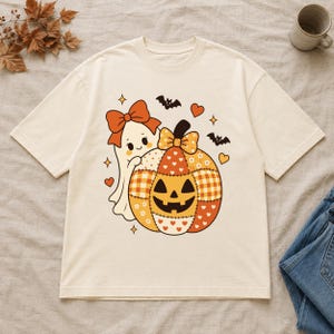 May include: A cream-coloured t-shirt featuring a graphic print of a white ghost wearing a large orange bow and a patchwork pumpkin with a matching orange bow. The design includes small black bats, orange hearts, and yellow star accents on the front.