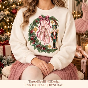 May include: A cream-colored crewneck sweatshirt featuring a graphic print of a Christmas wreath adorned with red berries, pinecones, and gold stars. Centered within the wreath are pink ballet slippers with ribbons and a nutcracker figure.