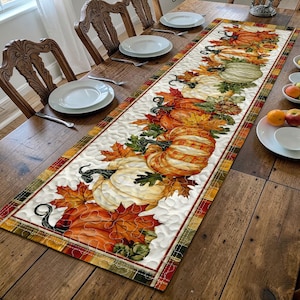 May include: A rectangular quilted table runner featuring a printed pattern of orange, white, and green pumpkins interspersed with colourful autumn maple leaves. The runner has a plaid border in shades of brown, orange, and green with wavy stitching.