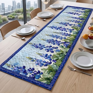 May include: A rectangular quilted table runner featuring a vibrant print of blue lupine flowers with green foliage against a light background, finished with a solid dark blue border and decorative white stitching throughout the fabric surface.
