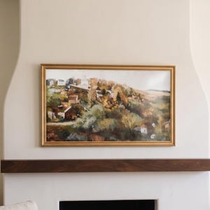 May include: A rectangular landscape painting in a gold frame depicting a hillside with trees in autumn colors, including shades of orange, yellow, and green, alongside small buildings nestled into the slope, mounted on a smooth white wall.