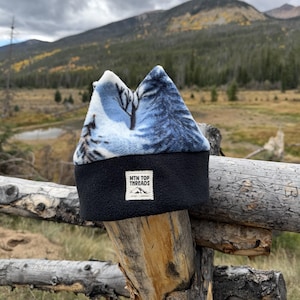 May include: A fleece beanie featuring a light blue and white winter forest print on the upper section and a solid black band on the lower section. A white rectangular patch on the front displays the text MTN TOP THREADS with a mountain icon.