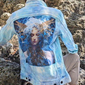Puede incluir: Una chaqueta vaquera azul claro con un patrón tie-dye. La parte posterior de la chaqueta presenta una ilustración colorida de una mujer con alas y una corona de plumas.