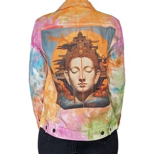 Puede incluir: Una chaqueta vaquera teñida con un diseño de tie-dye con una imagen colorida de un Buda en la espalda. La imagen presenta una ilustración detallada de un Buda con una expresión serena, rodeado de patrones intrincados y un halo dorado.