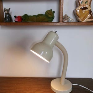 Puede incluir: Una lámpara de escritorio de color beige con una pantalla redondeada y un cuello flexible y acanalado. La lámpara cuenta con una base circular y un cable de alimentación blanco que sale por la parte trasera. La luz está encendida.