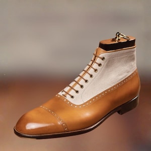 May include: A tan leather and cream canvas lace-up boot featuring a cap toe with decorative brogue detailing, thin brown laces, and a dark wooden shoe tree insert with a metal ring handle at the top, set against a neutral blurred background.