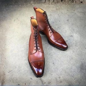 May include: A pair of polished tan leather lace-up boots featuring a cap toe design with dark brown burnished detailing at the front. The boots have a high ankle silhouette, dark brown laces, and a slim black sole with a small heel.