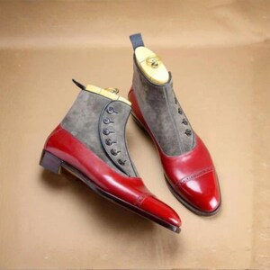 May include: A pair of ankle boots featuring a glossy red leather lower section and a grey suede upper shaft. The boots have a side button closure with five dark buttons, a cap toe with decorative brogue detailing, and a wooden shoe tree insert.